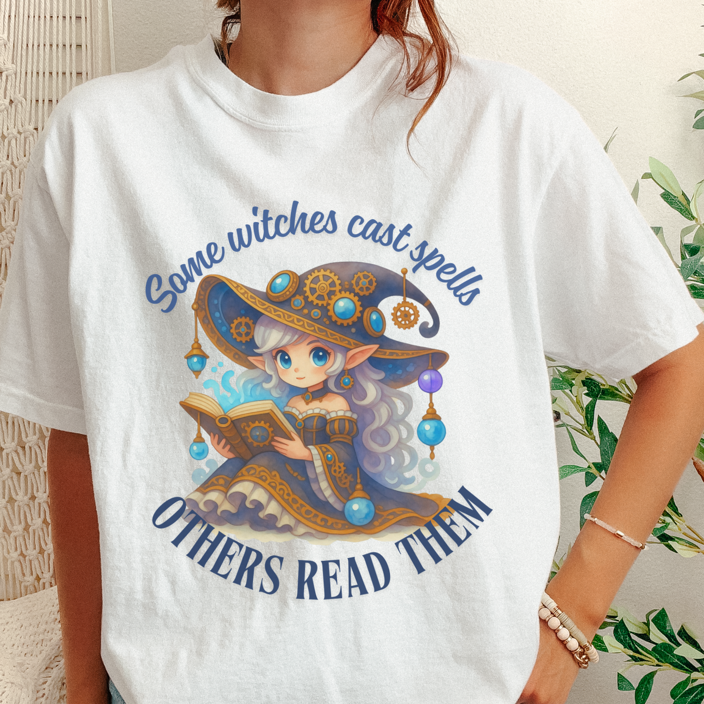 A person wearing a white graphic T-shirt with a colorful illustration of an elf-like witch in a large hat reading a book, surrounded by the text 'Some witches cast spells' and 'Others read them.' The photo is cropped from the neck to the hips, with a woven chair and green plant visible in the background.