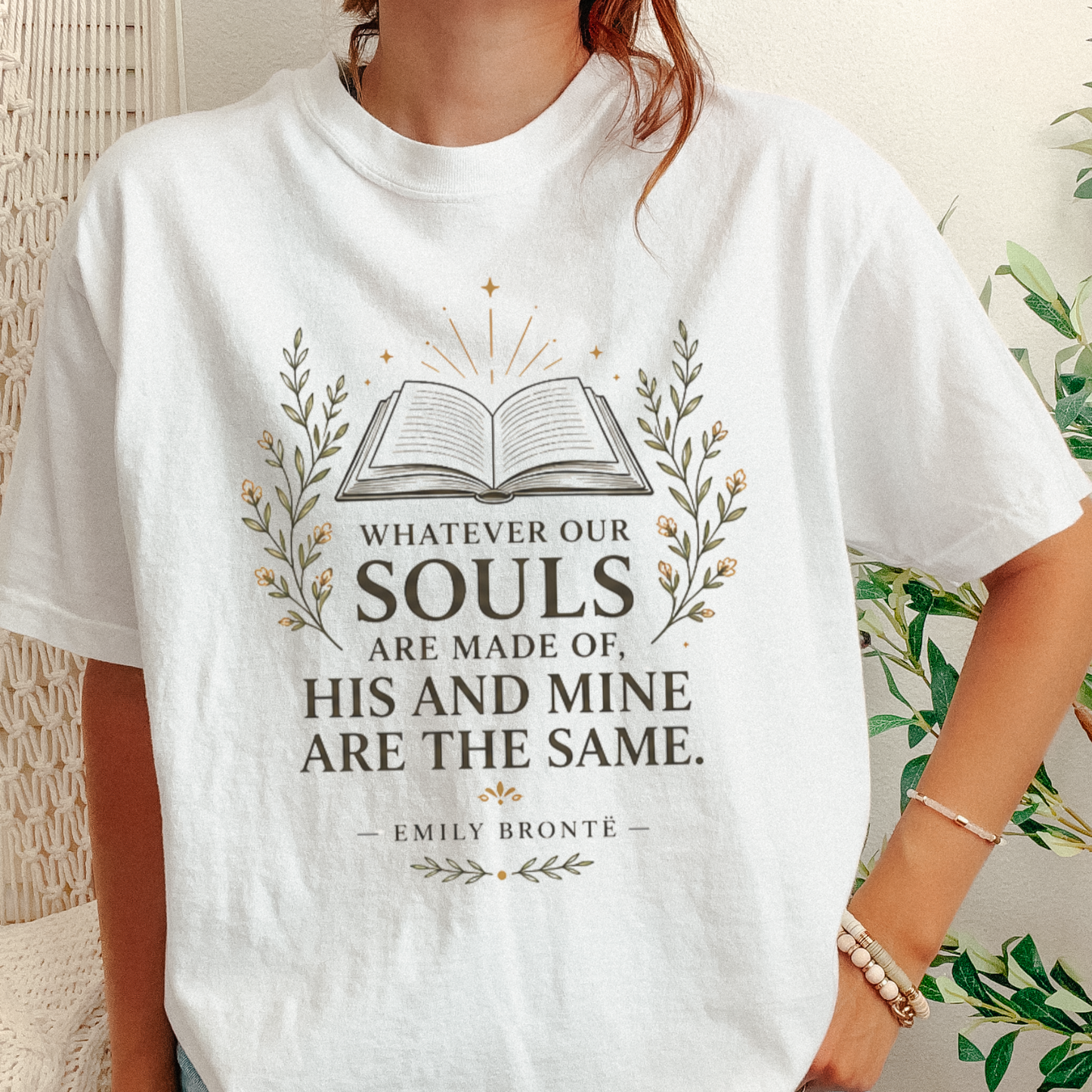 A woman wearing a white graphic t-shirt with a black printed quote by Emily Bronte: 'Whatever Our SOULS ARE MADE OF, HIS AND MINE ARE THE SAME.' The design features an open book surrounded by delicate floral illustrations.