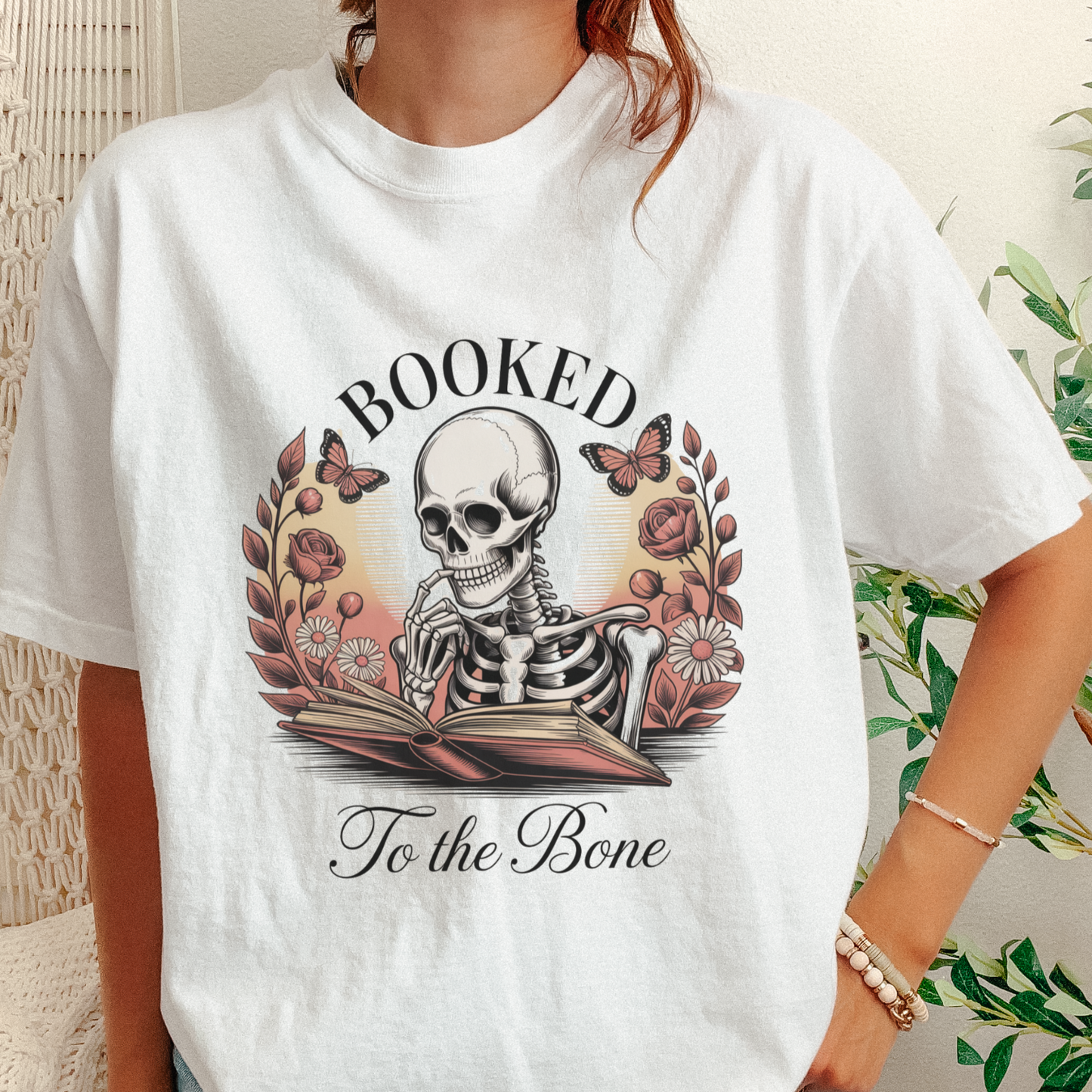 A person wearing a white graphic T-shirt with a large vintage-style print of a skeleton reading an open book, surrounded by flowers, butterflies, and the words “BOOKED To the Bone.”.