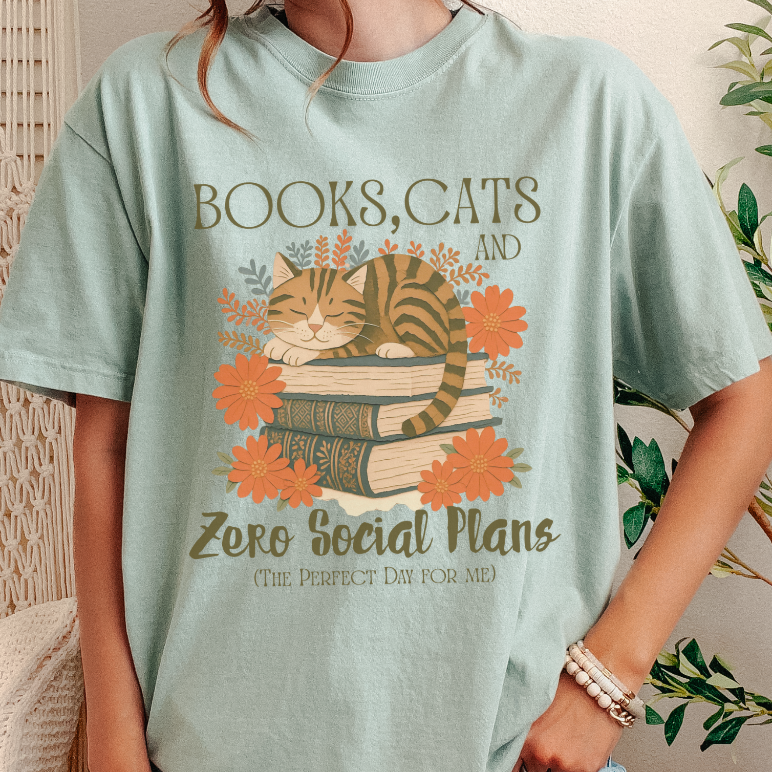 A person wearing a light mint green graphic t-shirt with a design of a cat sleeping on books surrounded by orange flowers and the text 'BOOKS, CATS AND Zero Social Plans (THE PERFECT DAY FOR ME)'.