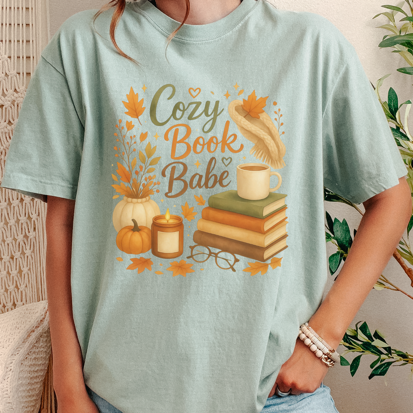 A person wearing a light mint green graphic t-shirt with a cozy autumn book theme, paired with light blue jeans in a home setting.