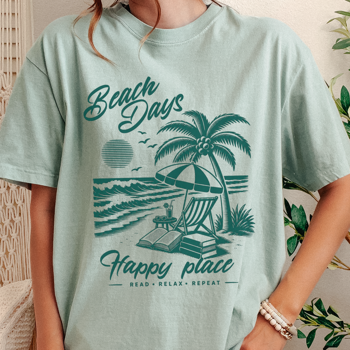 A person wearing an oversized light mint green graphic T-shirt with a teal beach scene print reading 'Beach Days Happy place' and 'READ RELAX REPEAT,' shown in a styled indoor setting with plants and woven decor.