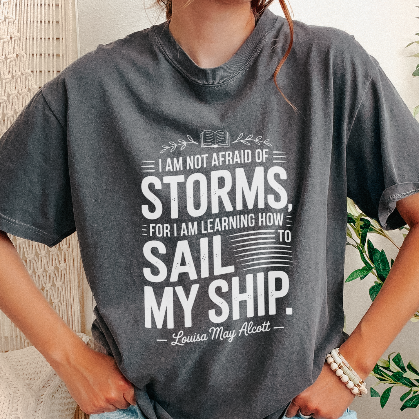A person wearing a dark gray oversized graphic T-shirt with white text that reads, 'I AM NOT AFRAID OF STORMS, FOR I AM LEARNING HOW TO SAIL MY SHIP. Louisa May Alcott.