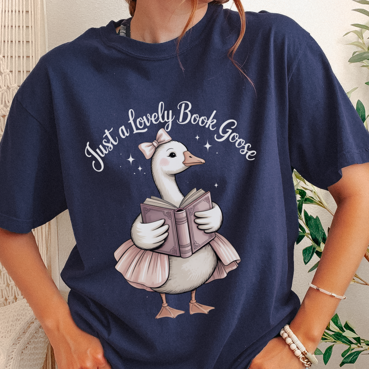 A person wearing a navy blue graphic T-shirt with a white goose illustration holding a book and the text 'Just a Lovely Book Goose' on the front, photographed indoors with a light neutral background and plant leaves visible.